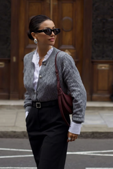 a lady in a ruffle collared shirt, a grey sweater and a pair of black pants with a belt